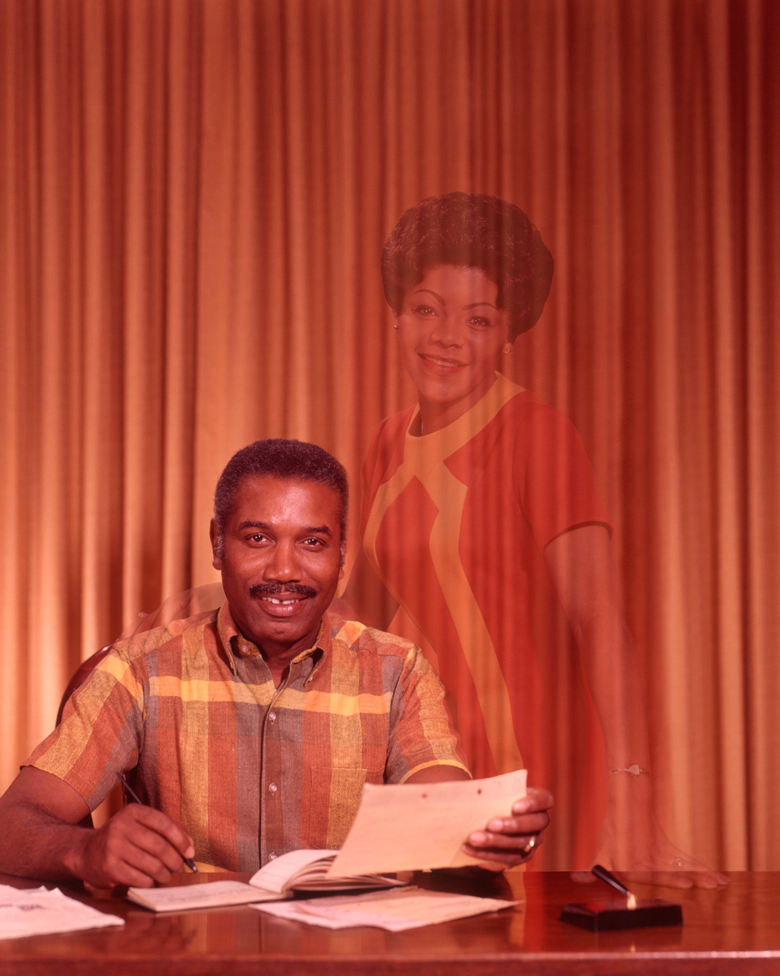 Colour photograph of a man sat on a desk with a pen and paper in hand. The photo has a hologram of a woman in view