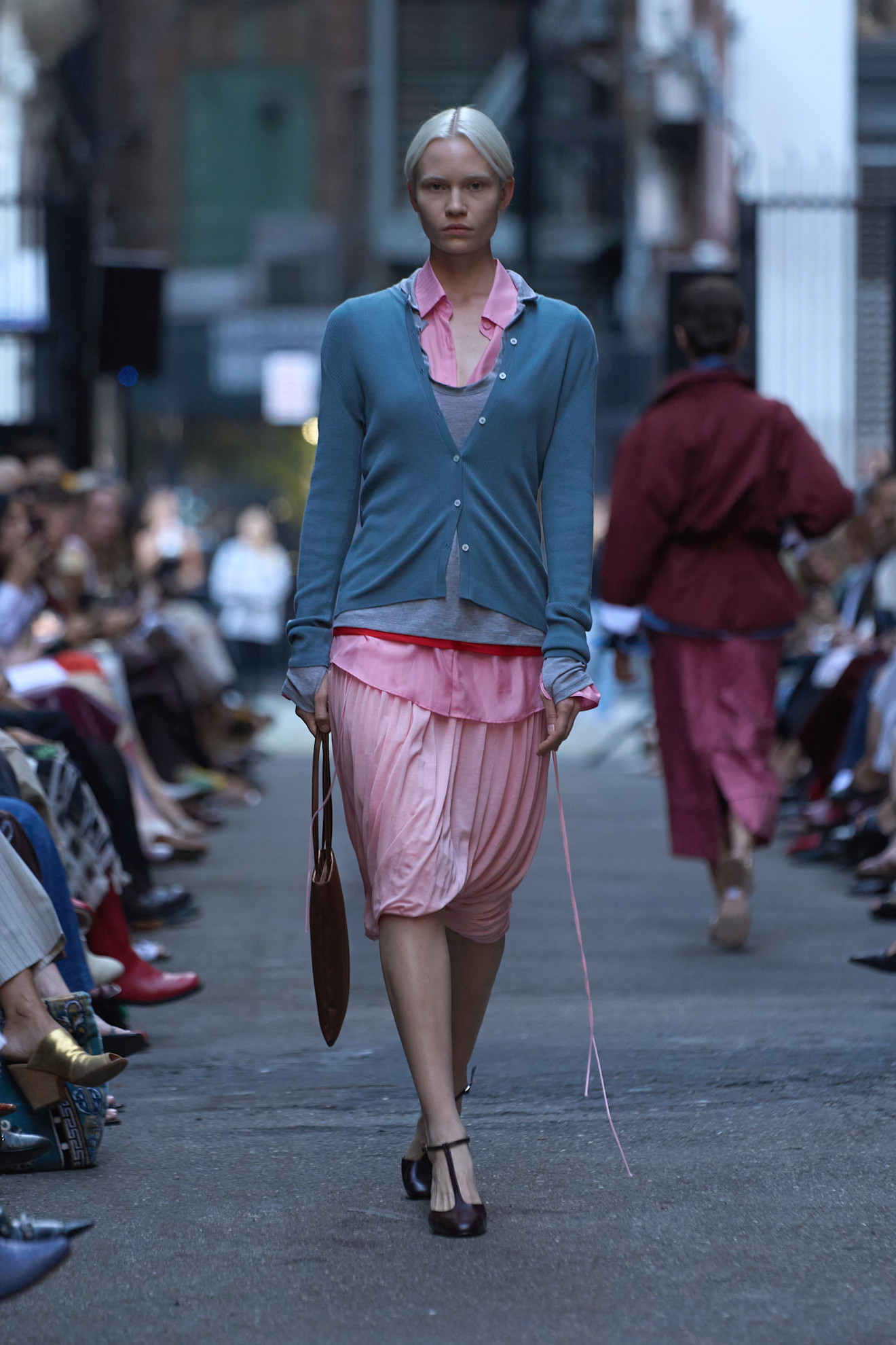Layering staples for spring 2026 is shown in a photo from Rachel Comey's runway show, with a model walking while wearing a pink button-down shirt layered under a gray and red tank top with a light gray cardigan layered on top. The layers are styled with a pink draped skirt, a red handbag, and black t-strap pumps 
