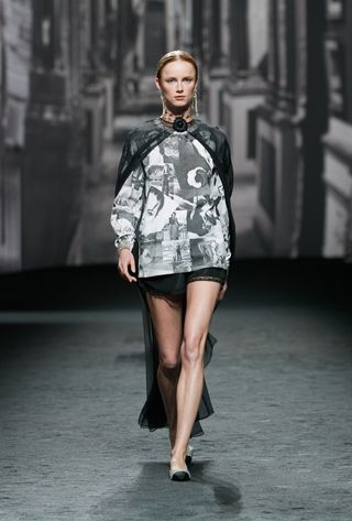 Chanel S/S 2023. A female model wearing a long sleeved shirt with black and white photos on it. a short black skirt, shiny pumps and a black lace cape.