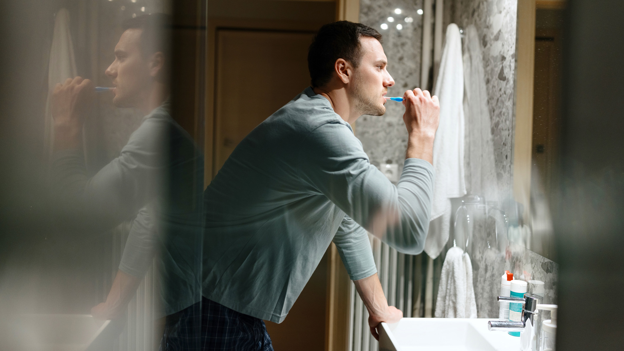 A dark haired man in a blue shirt brushes his teeth before going to bed