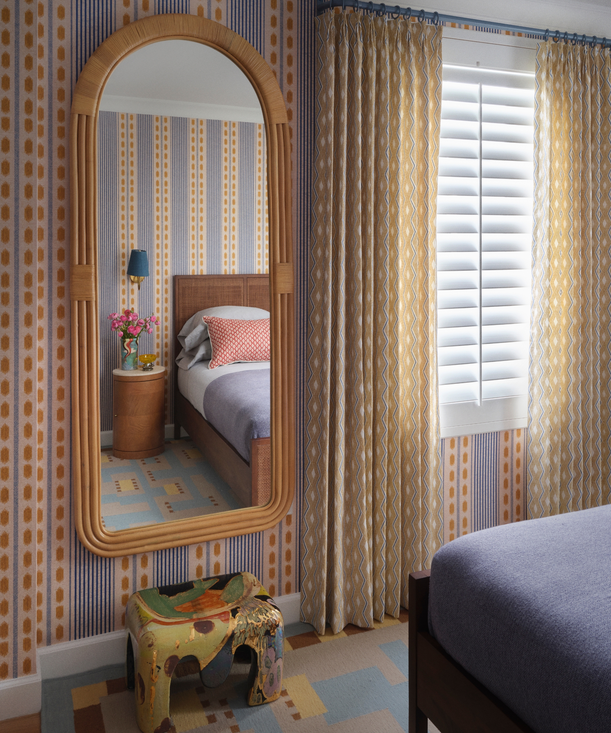 Guest bedroom with blue and ochre striped wallpaper, patterned curtains, rattan mirror, geometric rug and colorful ceramic stool.