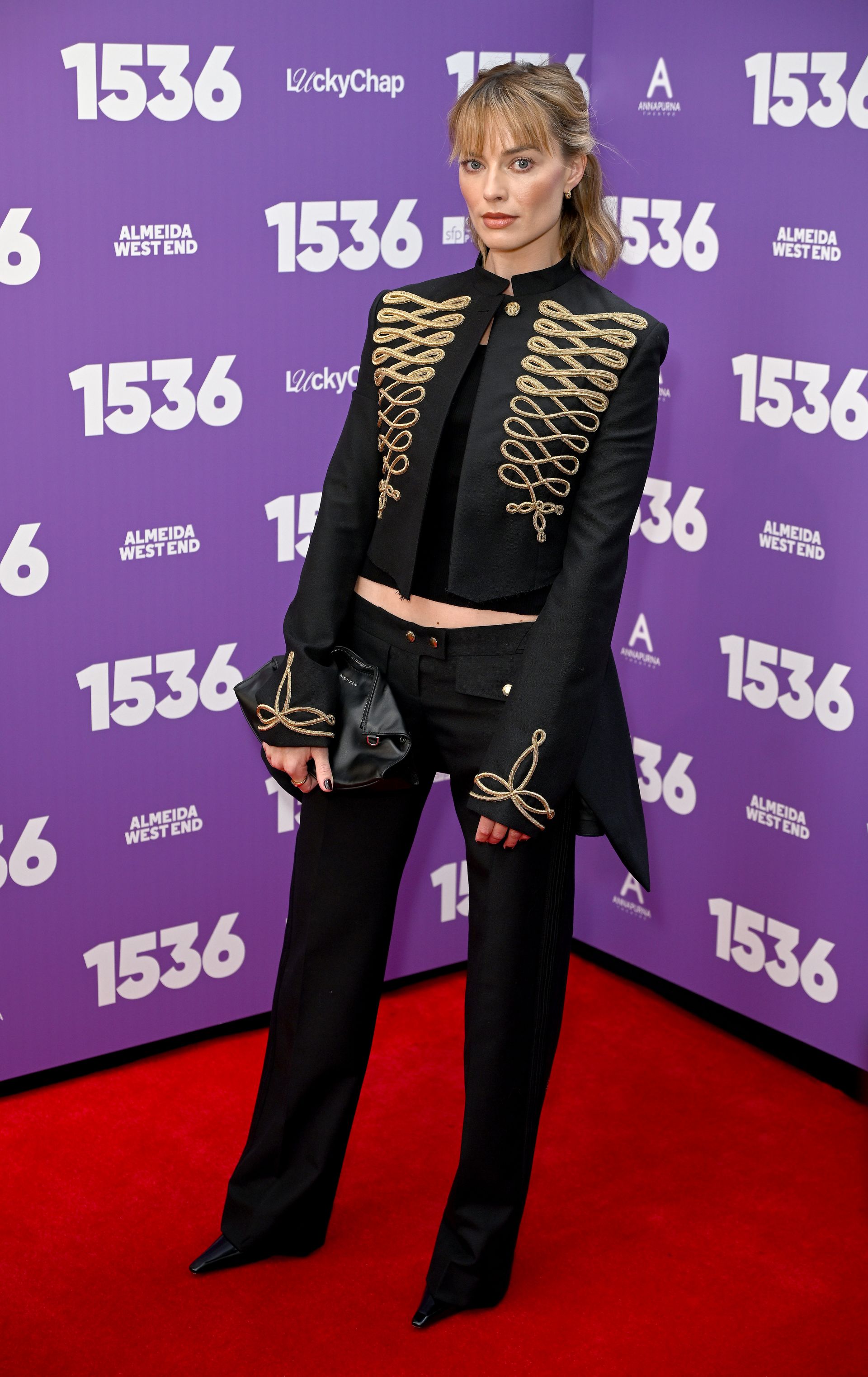 Margot Robbie wears a military jacket, low-rise black trousers, and a black clutch for the 1536 West End opening night in London.