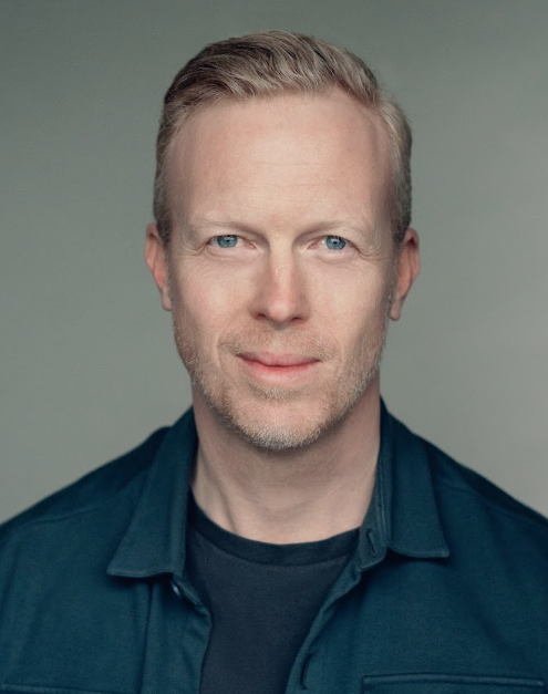 headshot of a man facing the camera wearing a dark shirt and tshirt underneath