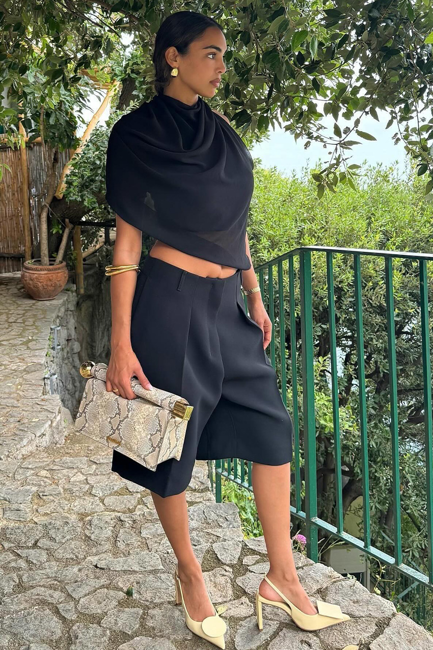 European vacation outfit idea is shown in a photo of a woman standing on a terrance wearing gold earrings, a black draped top, black bermuda shorts, gold bangles, a snakeskin clutch and yellow slingback heels