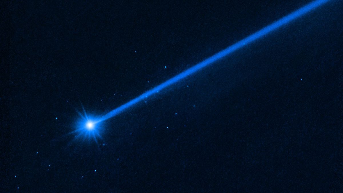 A Hubble telescope image of a bright blue asteroid, trailed by a long blue tail to the right. Small blue dots show boulders blasted away by NASA&#039;s DART mission.