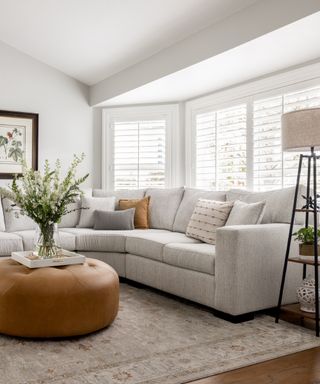 living room with white walls, shutters, pale gray sectional, floor lamp with storage,tan leather footstool, artwork, vase of flowers, neutral scheme,