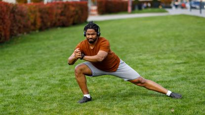 A man performs a lateral lunge outside on grass. His right knee is bent and his left leg is extended, his torso low to the ground. He wears headphones and clasps his hands in front.