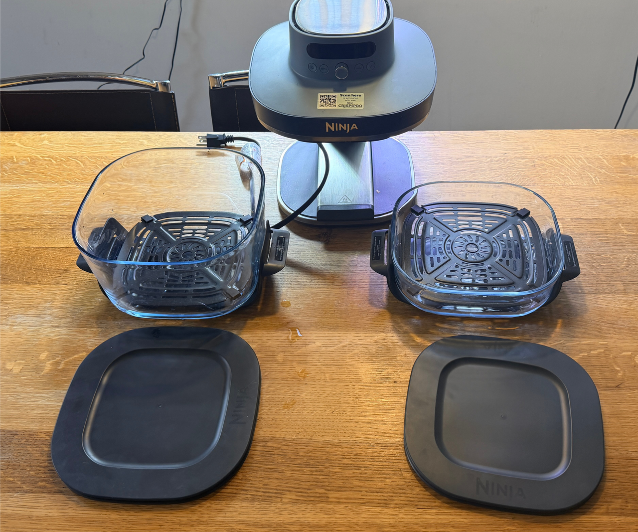 Two glass air fryer dishes beside one another with fitted crisper trays and marching plastic lids on a wooden counter, in front of the Ninja Crispi air fryer cooking head.