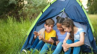 Tre barn med en mobiltelefon foran et telt i et grønt landskap.