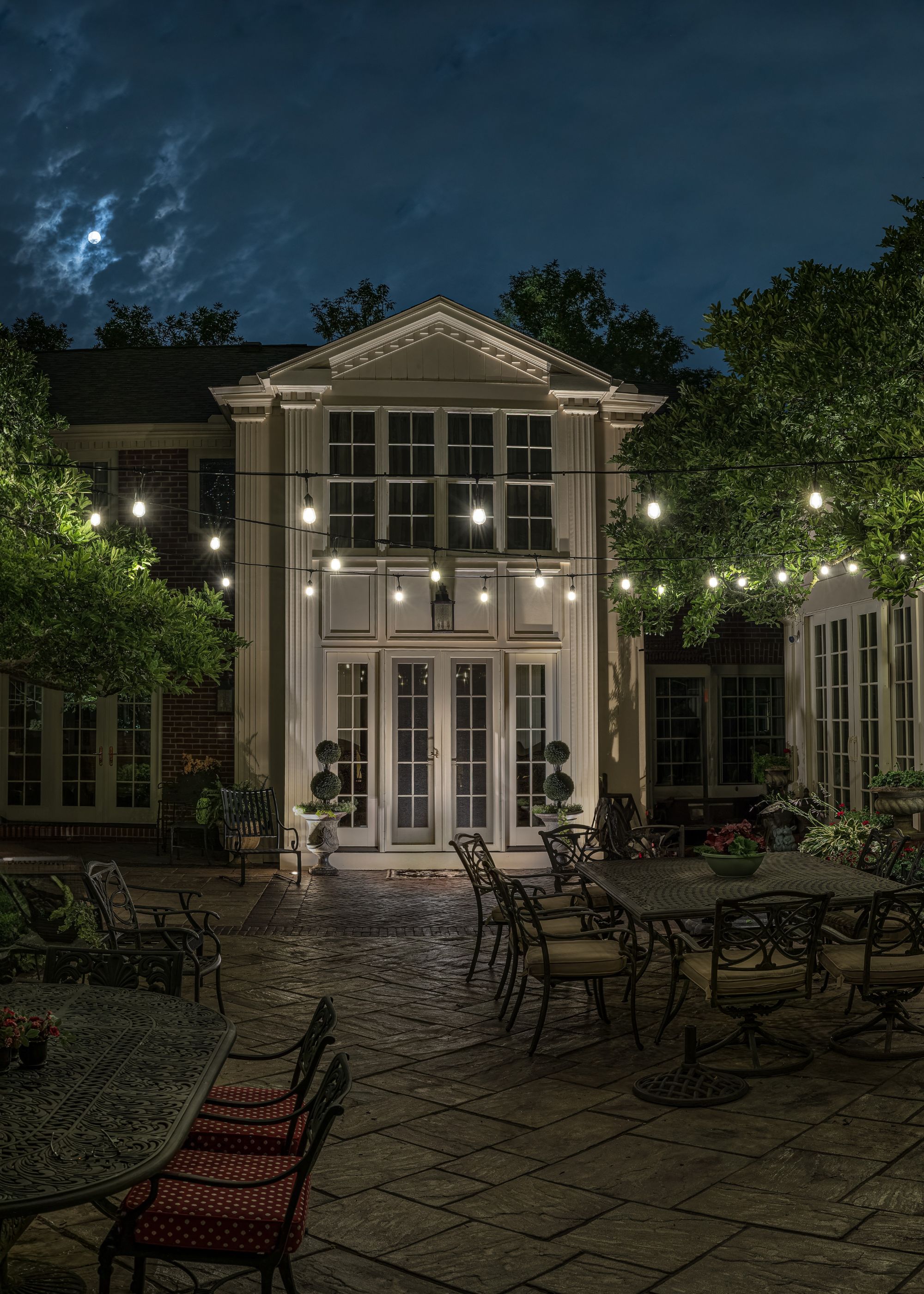 A large patio outside of a home with dining table and chairs. There are also string lights hanging above as well as uplighters near the trees and plants.