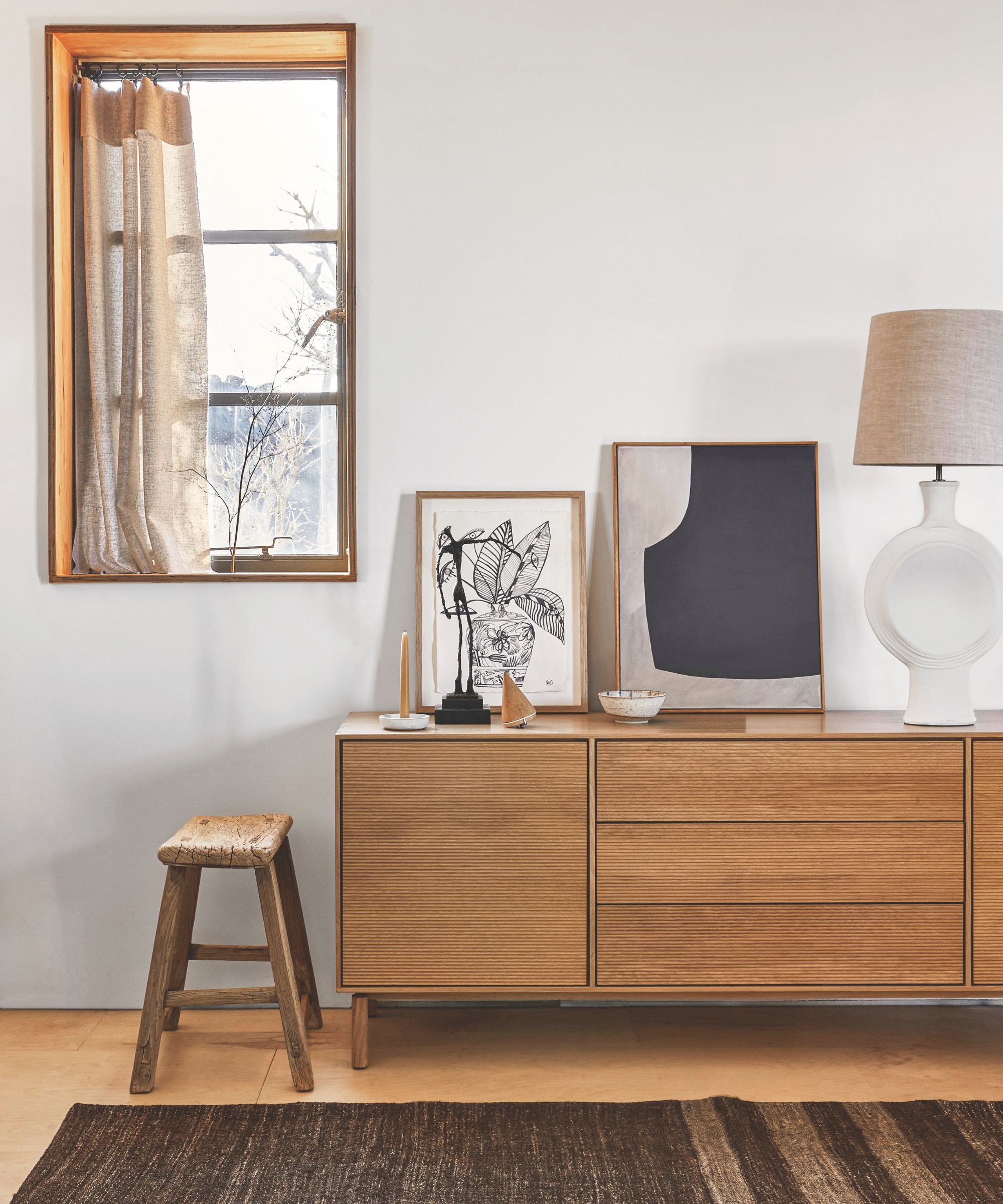 A neutral white room with wooden sideboard, window with beige curtains, artwork in wooden frames, white lamp with beige lampshade, wooden stool and striped dark brown rug.