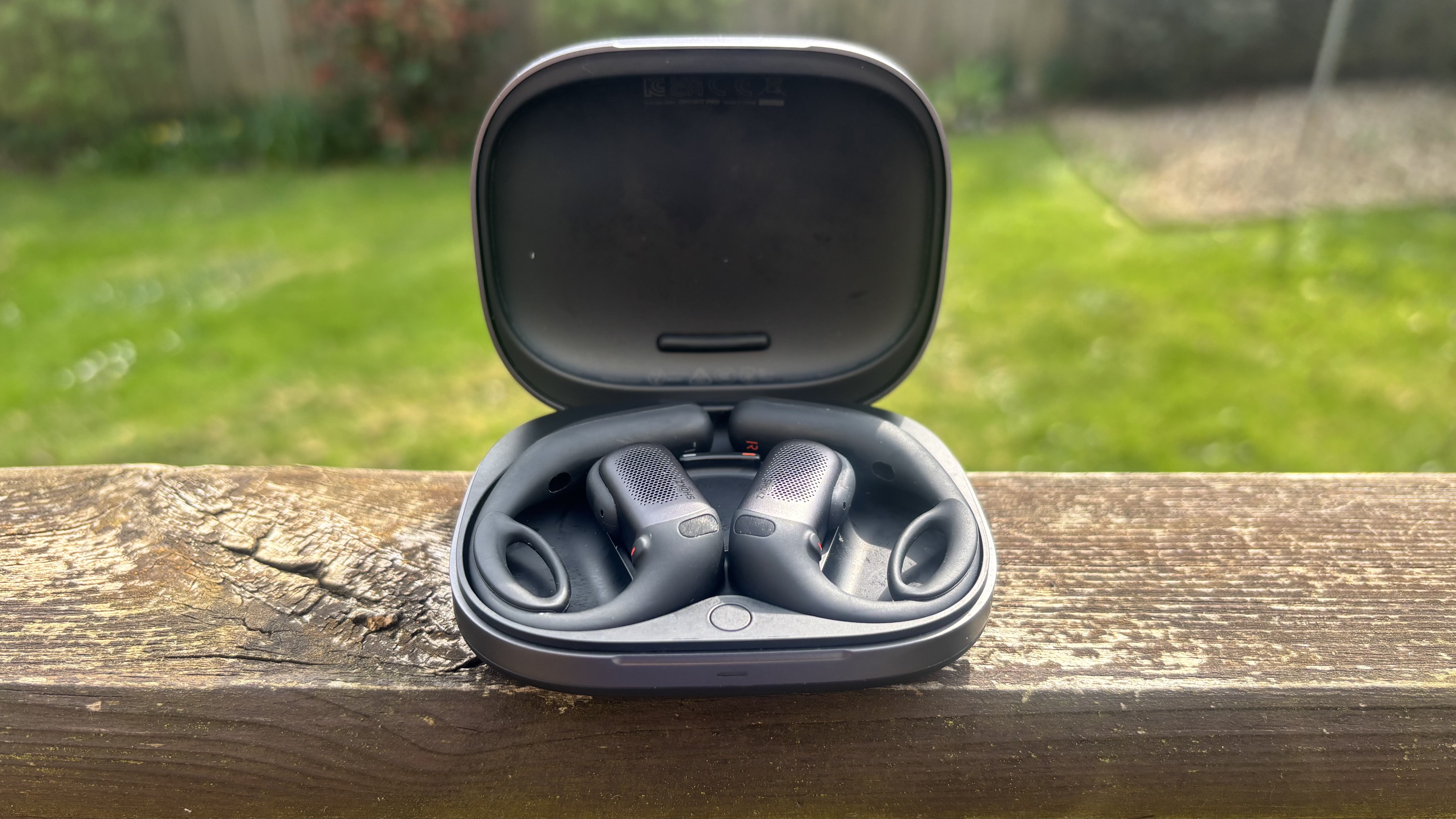 The Shokz OpenFit Pro earbuds, inside their charging case, on a wooden bannister with a grassy background.