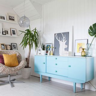Blue painted retro sideboard below shelves with photos on and patterned chair with yellow cushion