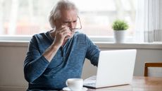 An older man looks concerned as he looks at his laptop while sitting at his kitchen table.