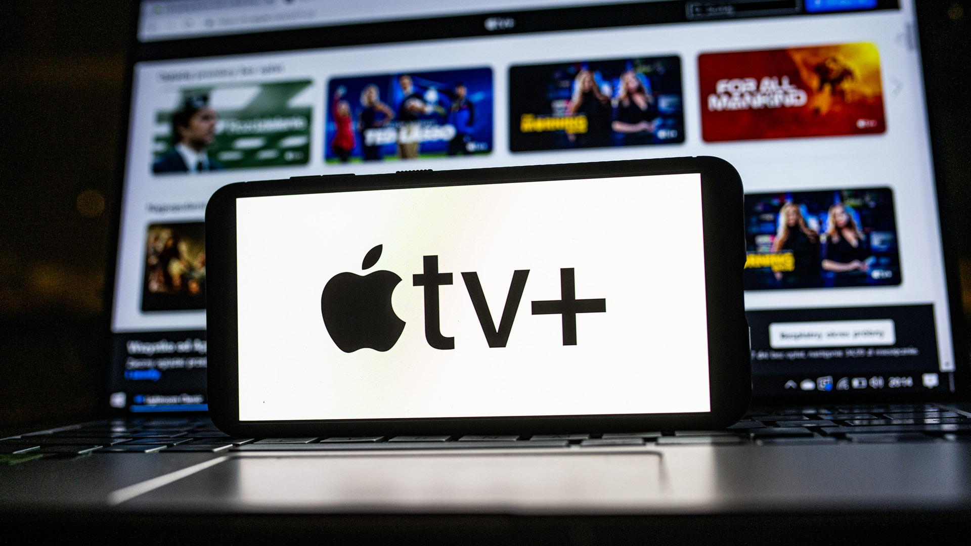 نشان‌واره Apple TV Plus روی تلفن همراهی که روی لپ‌تاپ نشسته است و چند نسخه اصلی Apple TV را نشان می‌دهد