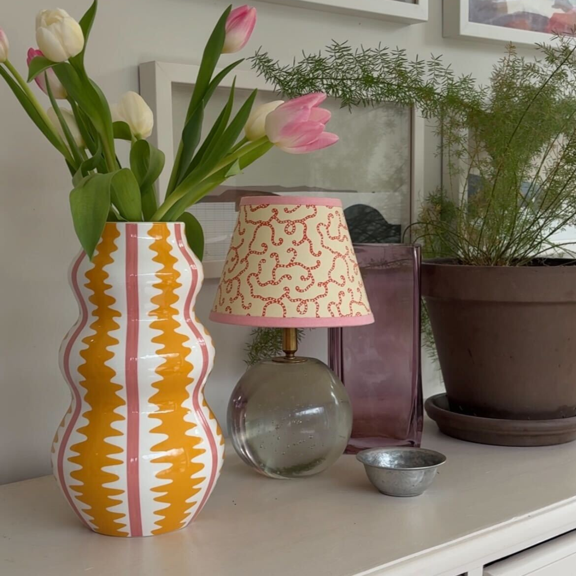 Habitat yellow and pink wiggle vase holding pink and white tulips on a white sideboard. Next to it is a glass lamp with white and pink shade, and a houseplant.