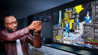 Masc presenting person holding G'AIM'E light gun in front of large screen featuring Time Crisis gameplay.