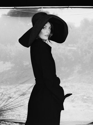 Leila George wears black floppy hat with black belted coat.