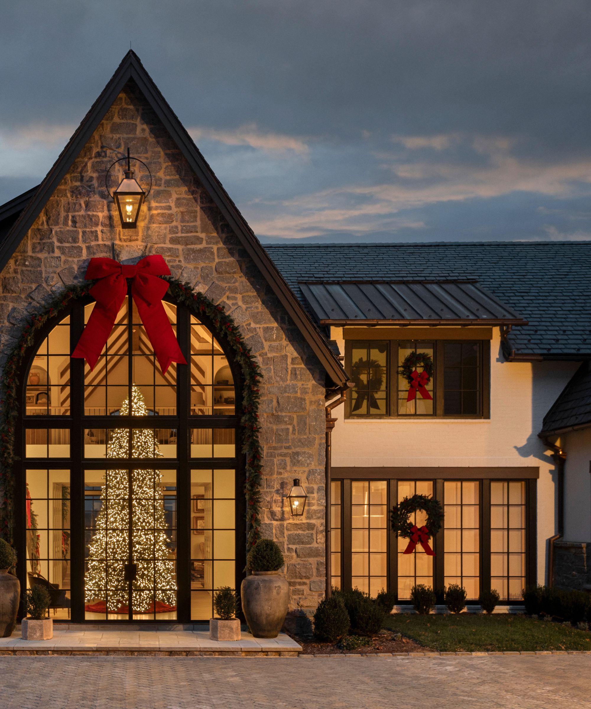 tall stone home with a pitched steep roof with a red bow, glass door with a tall lit tree and wreaths with ribbons on the windows