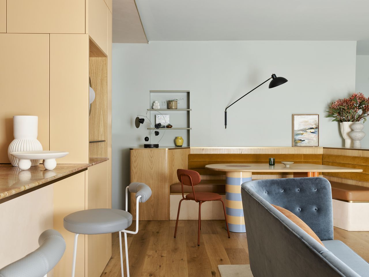 colorful dining zone with banquette seat, striped table with timber top, leather upholstery and timber frame. the walls are painted a soft light blue and have a niche with shelves and a reaching lamp. the kitchen joinery is painted a soft apricot with blue bar stools