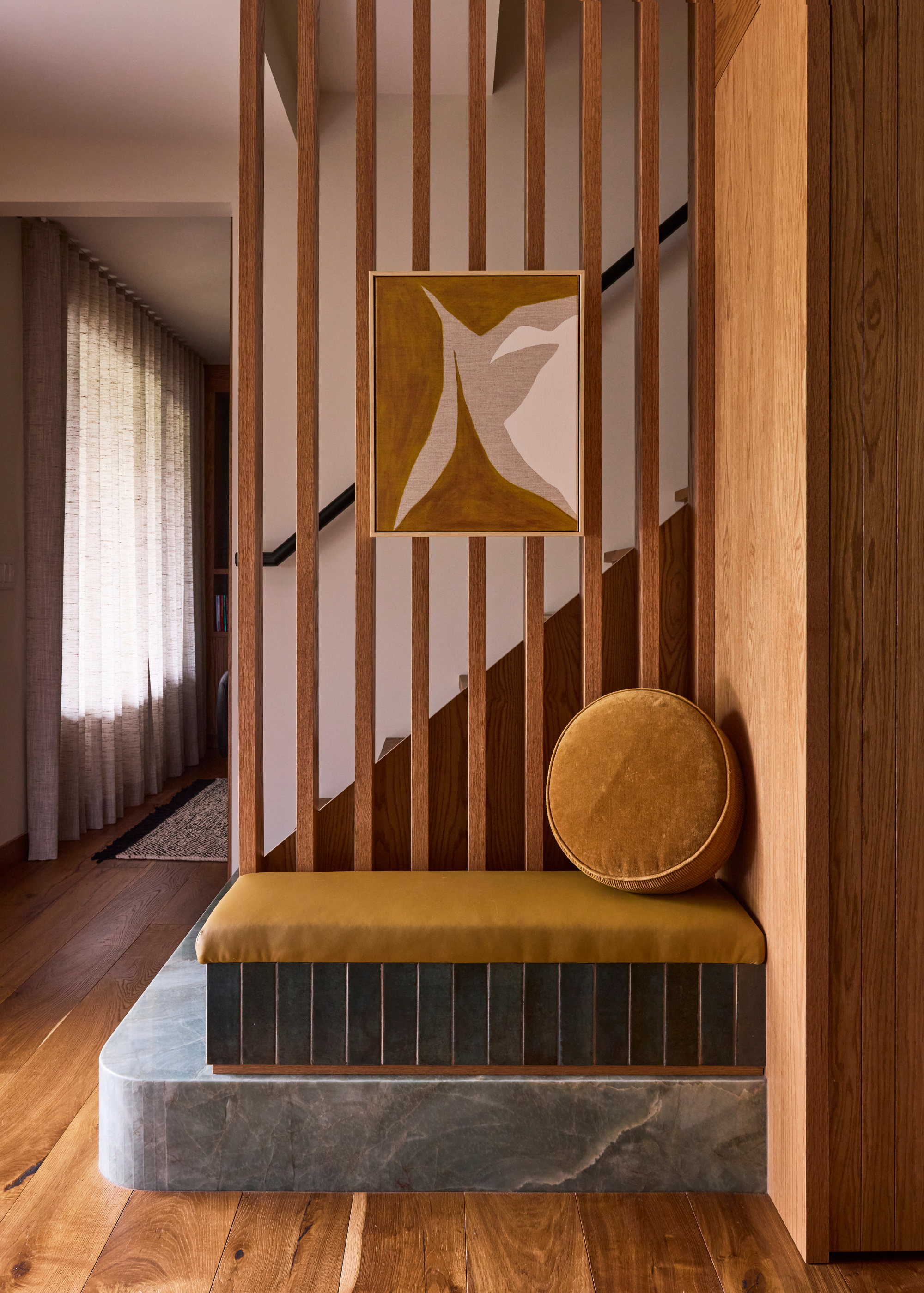 An entryway with a built-in bench with a green tile base and a mustard upholstered top and round cushion by wood stairs with a marble base
