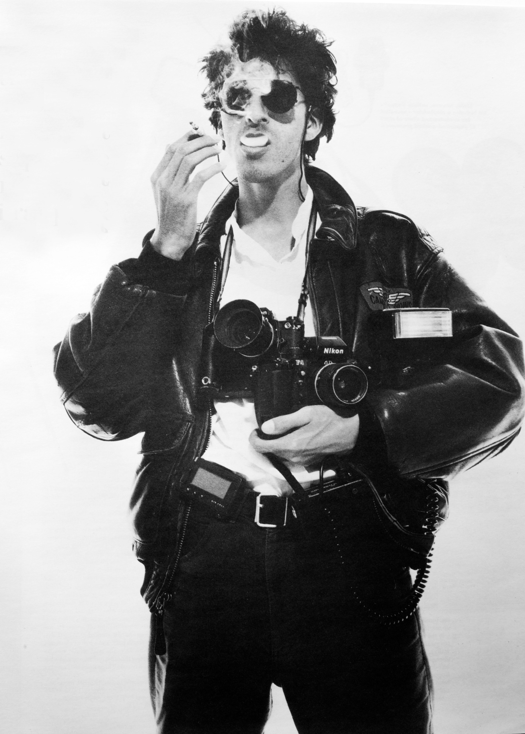 Black and white portrait of a young David Koppel in sunglasses and a leather flight jacket, exhaling cigarette smoke with two Nikon cameras, a flashgun and a pager strapped to his body.