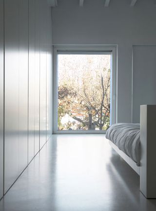 White bedroom, displaying the corner of a bed and showing the view from the window looking out onto a tree