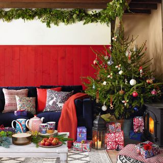 Decorating ideas for Christmas, foliage garlands on the ceiling, blue sofa in front of red painted panelling, cushions with floral patterns, stack of presents underneath a decorated Christmas tree