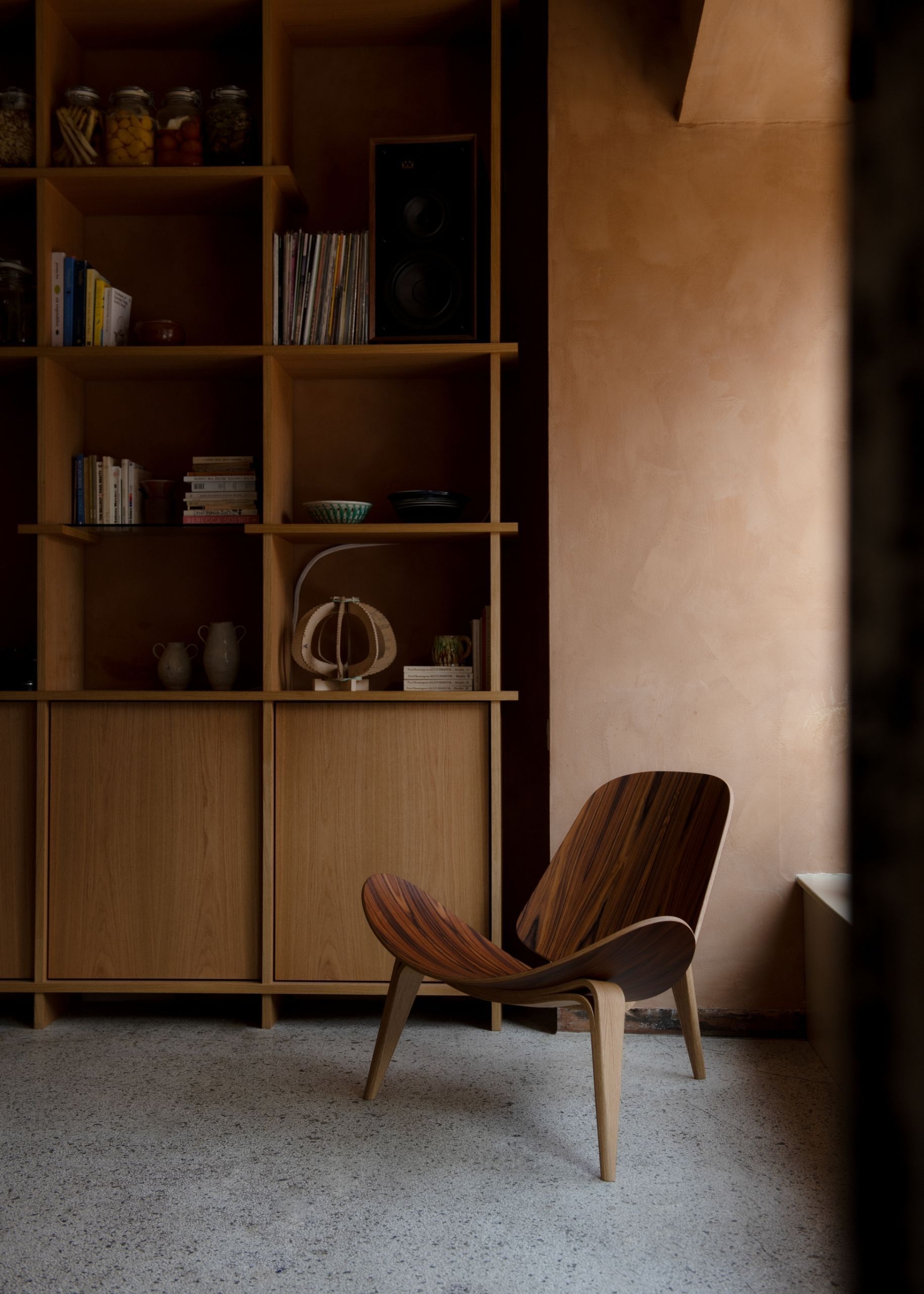 Image of a wooden shell chair in a room with terracotta walls, a wooden built-in bookcase, and terrazzo floors.
