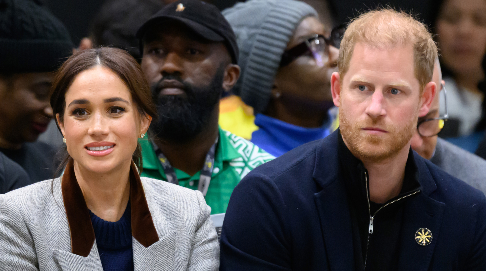 Meghan Markle wears a gray blazer and sits next to Prince Harry, who has balding red hair