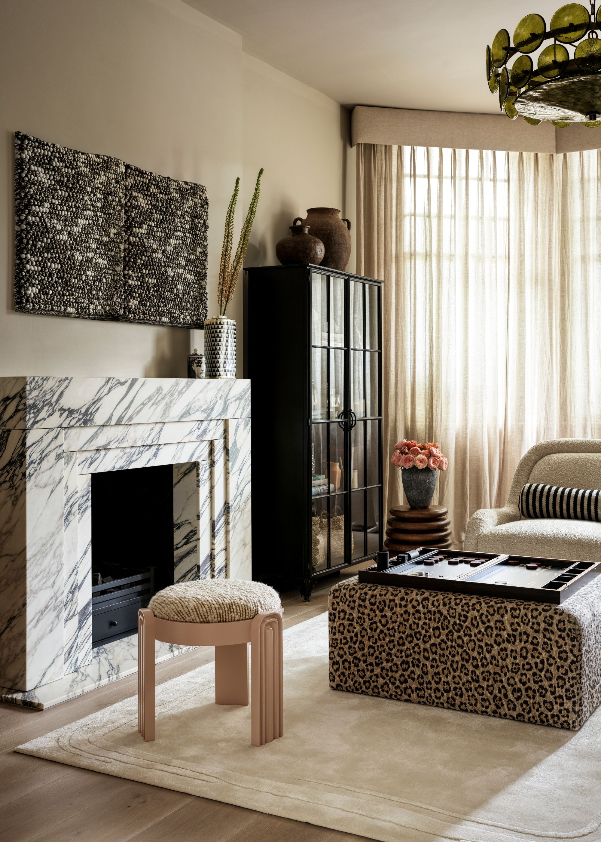 A living room with beige walls, a large cream rug, and a marble fireplace with a tapestry above it. A bay window with sheer curtains and a leopard print ottoman in the middle of the room.