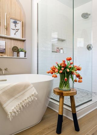 Warm neutral bathroom with roll top tub and tulips on a stool in front of screened shower