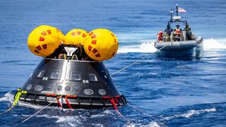 a cone-shaped spacecraft with inflatable balloons on top floats in the ocean. it is on a tow line behind a small boat that is pulling it across the water
