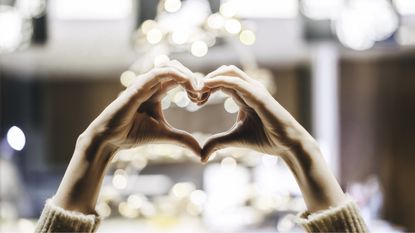 A woman forms a heart shape with her hands against the backdrop of white holiday lights.