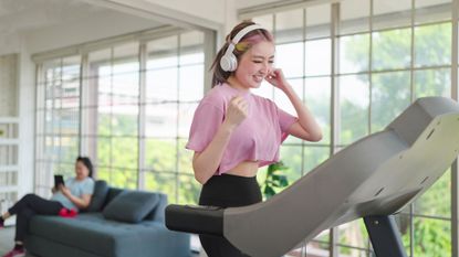 A smiling woman in a pink crop top and black leggings walks on a treadmill. She wears white over-head headphones and her arms are raised as though dancing.