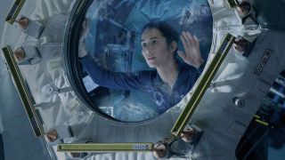 a woman in a blue flight suit looks through a glass hatch in a cramped laboratory filled with wires and screens