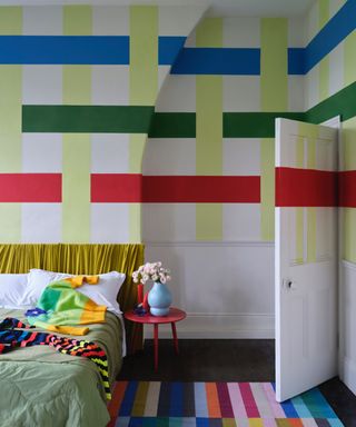 What is Bauhaus style, bedroom with bold paint effect check with lime, blue, green and red stripes, yellow bed, striped rug, red stool as bedside