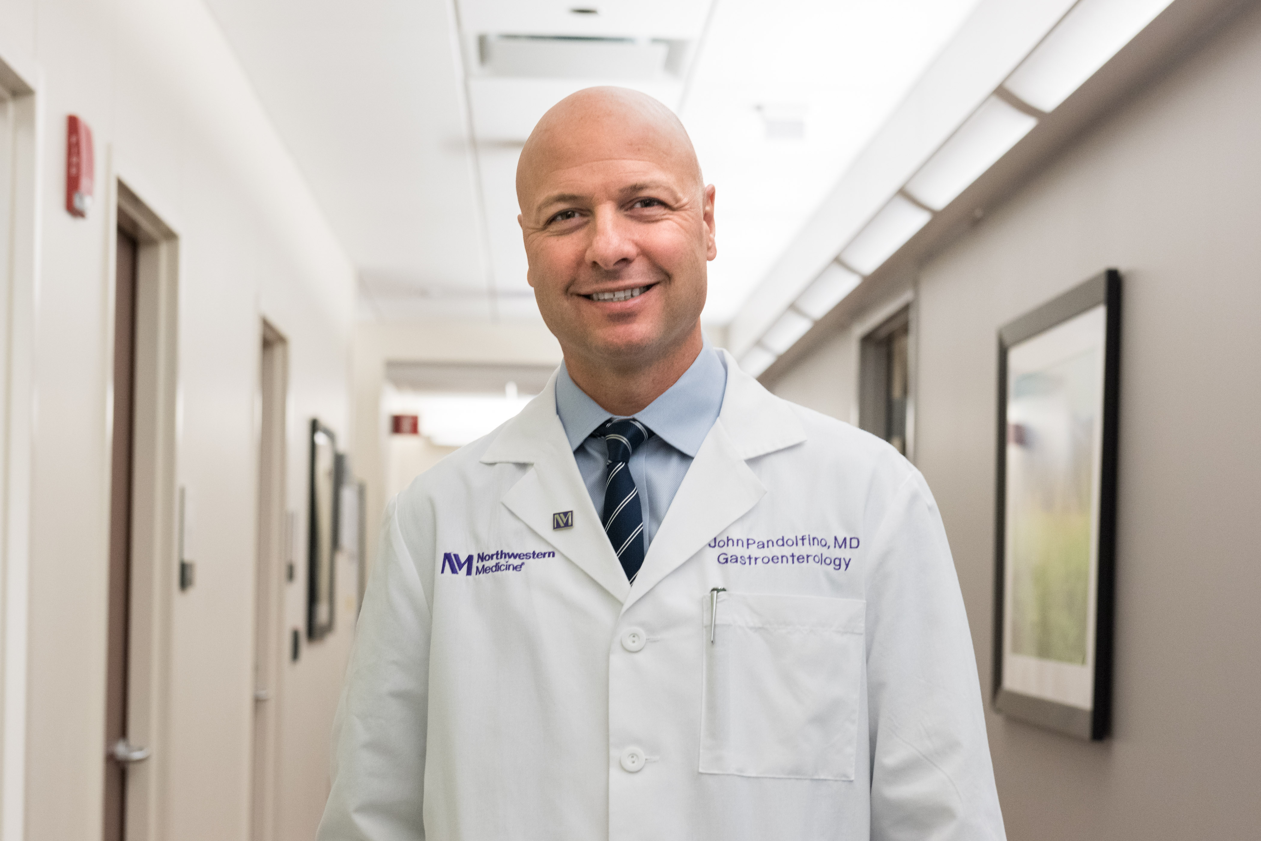 A bald man wearing a blue striped tie over a blue button down shirt with a lab coat over it stands in a hallway looking at the camera