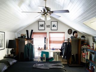 Attic bedroom interior, bed with black sheets, white desk with green swivel chair, ceiling fan, two windows with red blinds, framed artwork on white walls, dark grey floor, book case to the right, leather jackets and shirts hung on clothes rails, tall black leather boots, bedside table with black lamp, white wooden slat ceiling, police caps