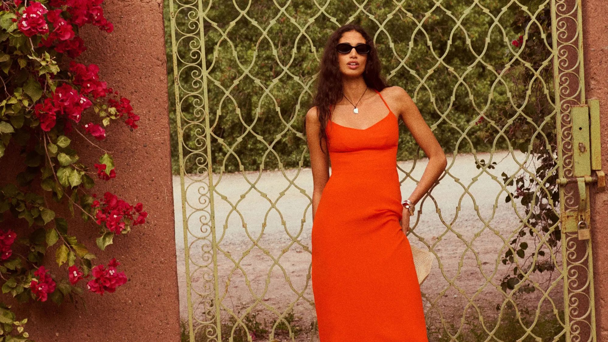 a model poses in front of a metal gate in an orange dress and sunglasses