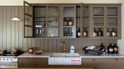 Taupe kitchen cabinets with glass fronts, butler sink, brass fixtures, and paneled walls. There are amber glass jars and a cookbook in view