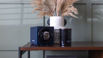 One of the best watch winders on a wooden table in front of a green wall, with a vase and candle next to it