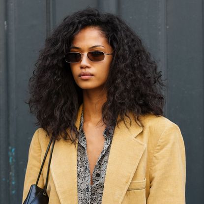 A woman with beautiful naturally curly hair wearing a beige blazer and sunglasses looks at the camera