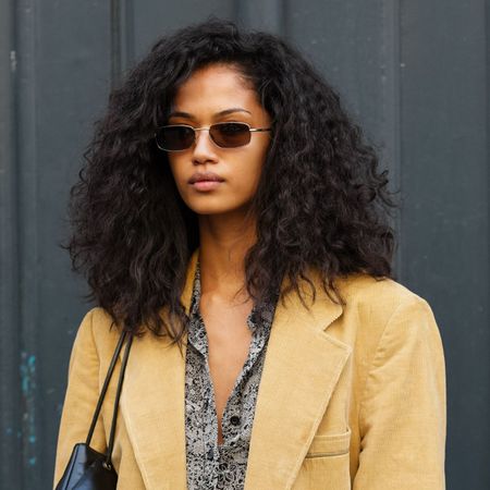 A woman with beautiful naturally curly hair wearing a beige blazer and sunglasses looks at the camera