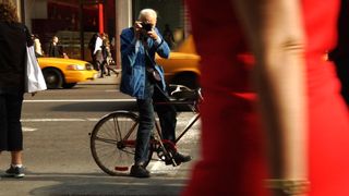 Bill Cunningham on bicycle taking a picture