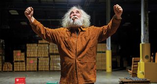 EHX founder Mike Matthews stands in a warehouse with his fists in the air. He wears a tan jacket and has a trademark cigar in his mouth.