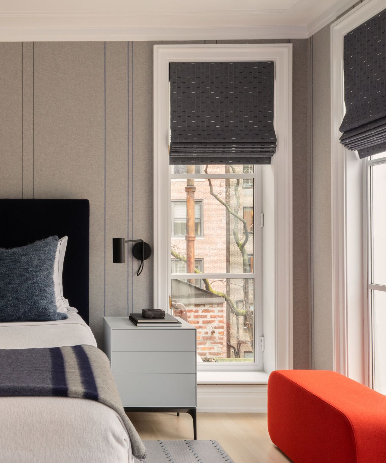 A bedroom with striped wallpaper, two large windows, and a bright red bench