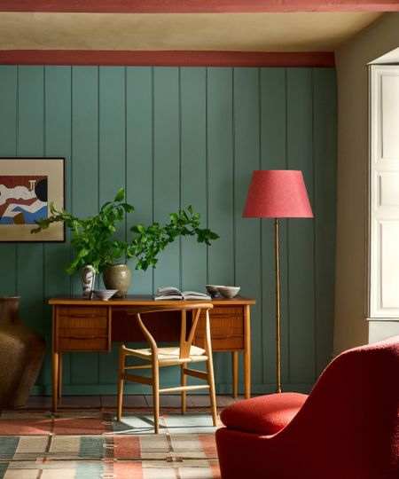 Living room painted in Little Greene's Perpetual Blue, a wooden desk and chair, a pink floor lamp and pink beams, and a geometric rug and red armchair.