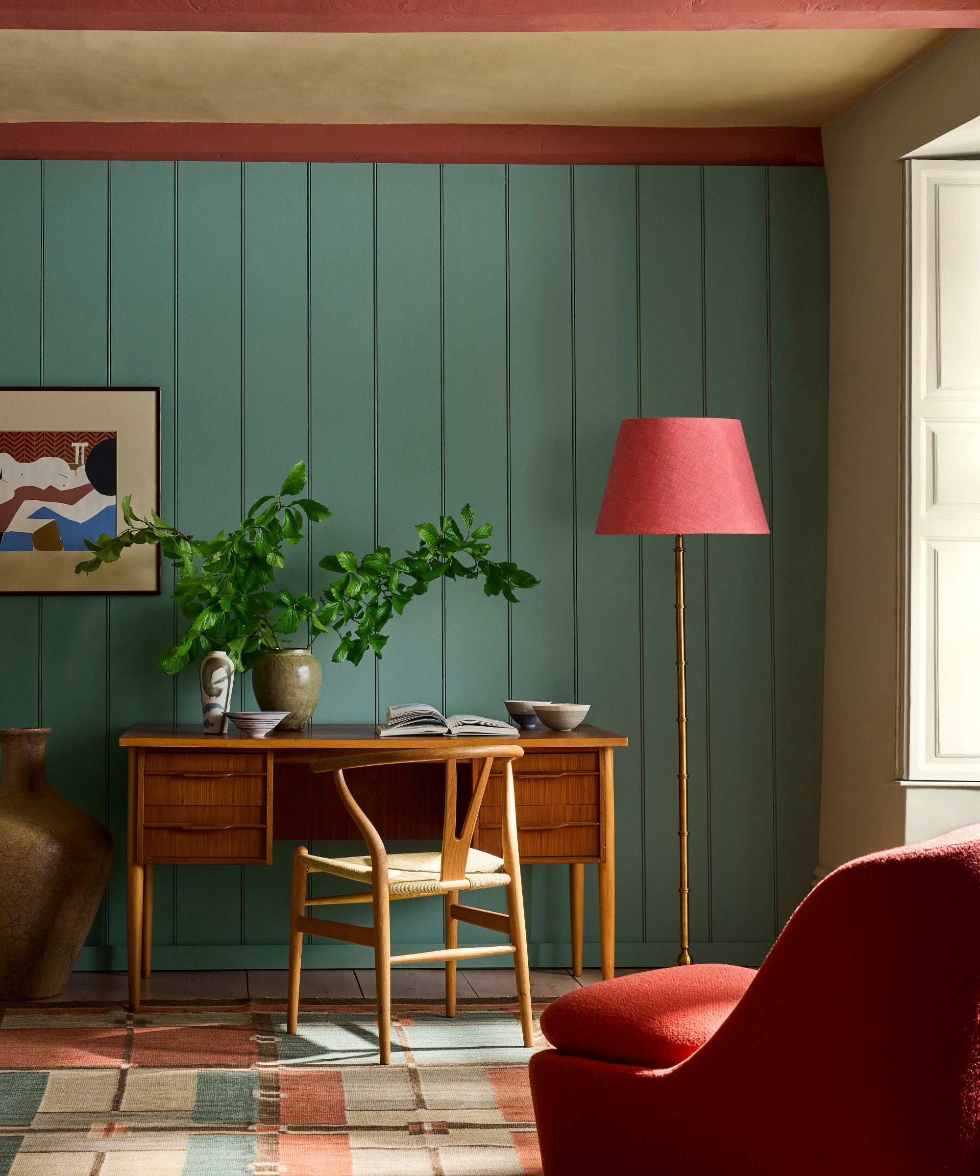 Living room painted in Little Greene's Perpetual Blue, a wooden desk and chair, a pink floor lamp and pink beams, and a geometric rug and red armchair.
