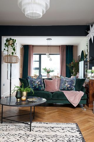 A living room with green velvet sofa and pink cushion decor with black and white rug on parquet floor